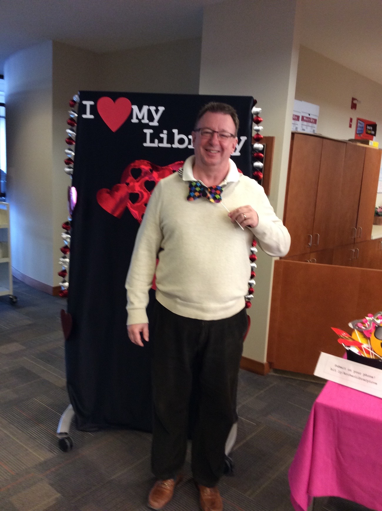 University Librarian Brett Waytuck stands in front of the I love my library backdrop holding up a prop polkadotted bowtie on a stick and smiling